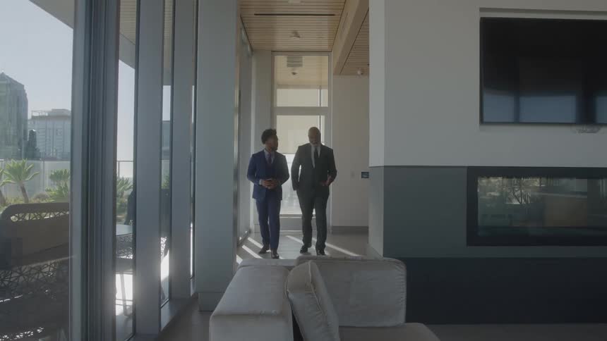 Two men in suits talking as they walk side by side on a hallway leading to the lobby of a building
