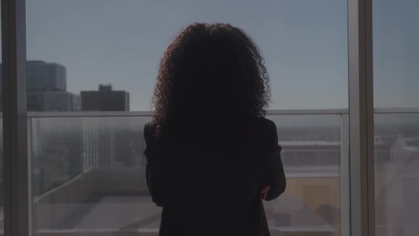 A wide view from behind shows a woman with curly hair and a black jacket standing in a skyscraper office with her arms crossed while looking out a large window at a city view on a sunny day