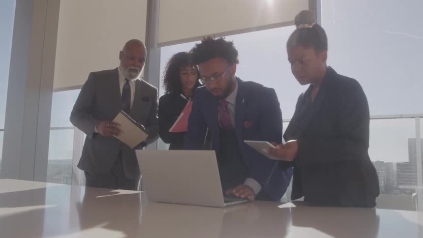 A team of four professional men and women stands around a conference table in an office looking at a laptop and working together with a cityscape visible through the windows
