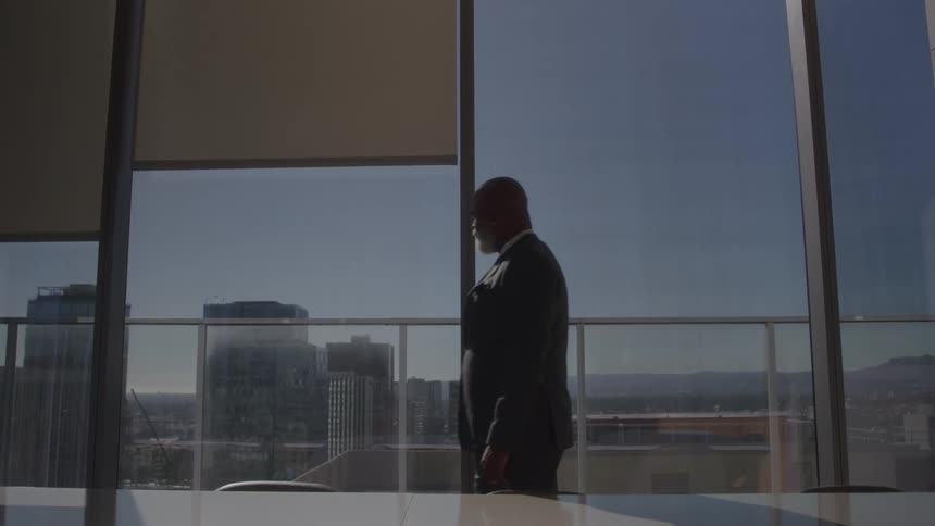 A man in a suit walks in an office and looks to the outside with a pensive expression