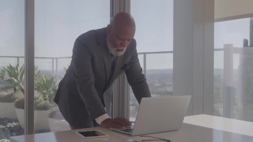 In a sunlit high rise office a young businessman in a suit and glasses stands and types on his laptop at a desk