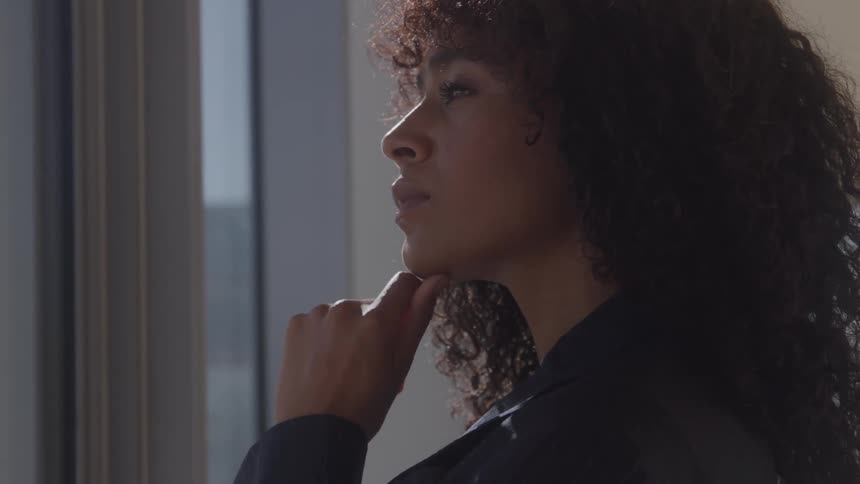 In a hazy backlit close up a pensive businesswoman with curly hair stands in profile thoughtfully resting her chin on her hand
