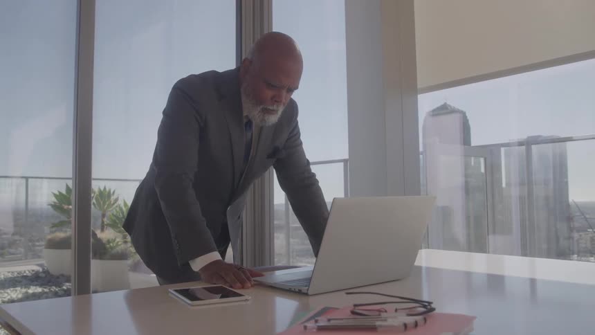 A man in a suit stands in an office and works on a laptop