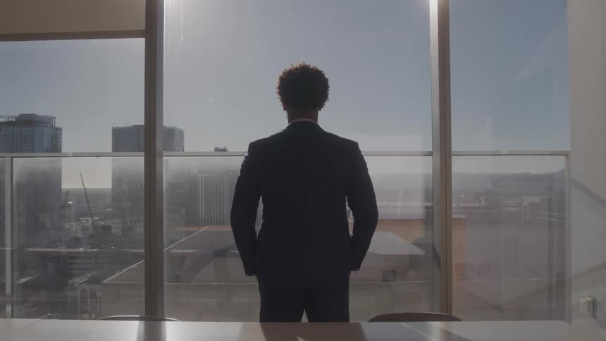A man in a suit stands and looks out a window at a city