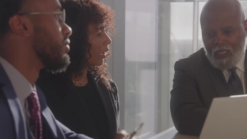 A team of two men and a woman in professional business attire is sitting at a conference table having a serious discussion while looking at a laptop