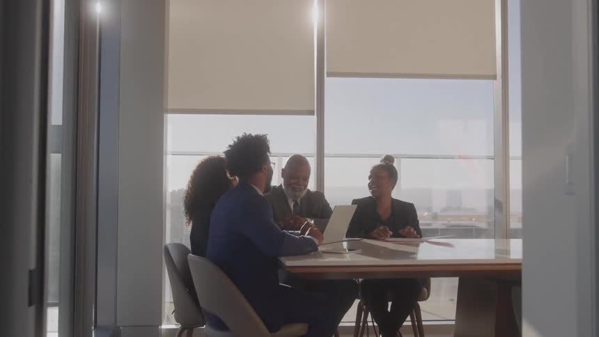 Two office workers giving a presentation to an elderly man and his wife all wearing suits seated in a conference room during a midday discussion