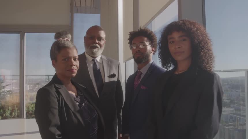 In a sunlit modern office a team of four diverse business professionals stands together posing confidently for a group portrait