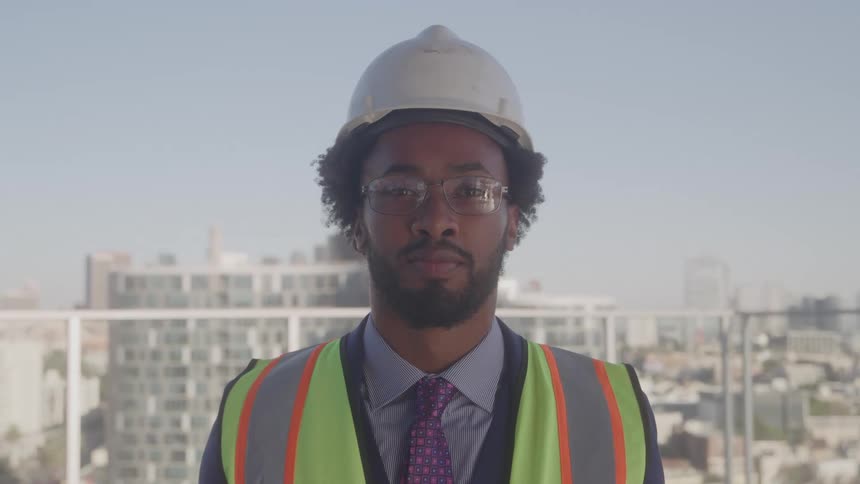 Black construction worker wearing a green safety vest hard hat and glasses standing inside a highrise building at midday with the city skyline visible in the background
