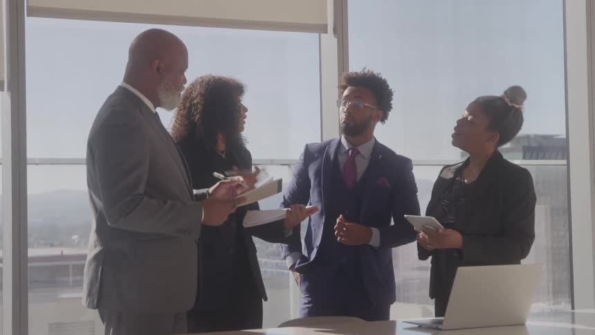 A group of four black executives discussing while standing at a table in a meeting room during daytime