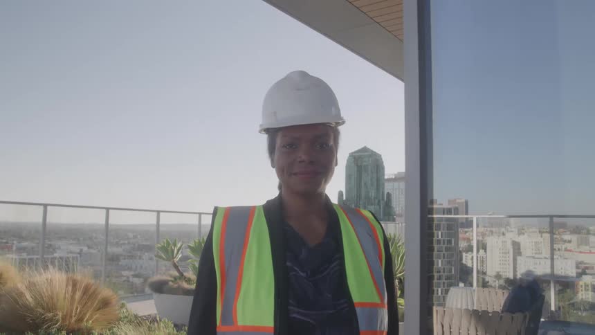 A woman wearing a hard hat and safety vest stands on a high rise balcony overlooking the city and smiles at the camera