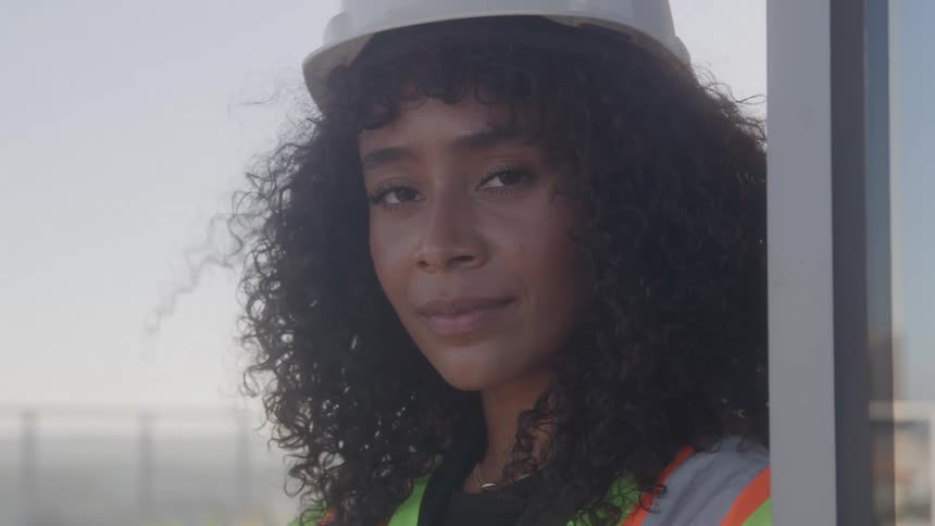 A woman wearing a hard hat and a reflective vest poses for the camera on a balcony