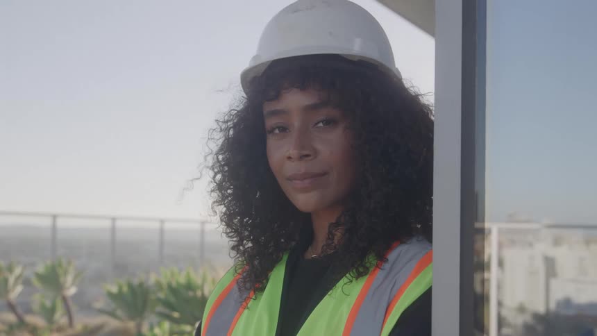 A woman wearing a hard hat and a reflective vest poses for the camera on a balcony