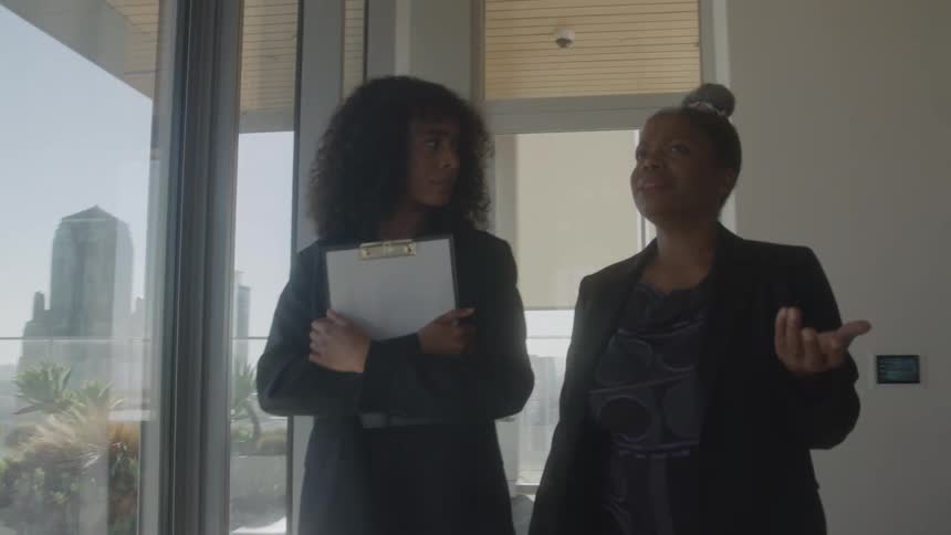 Two black female coworkers converse while walking together in an office during daytime