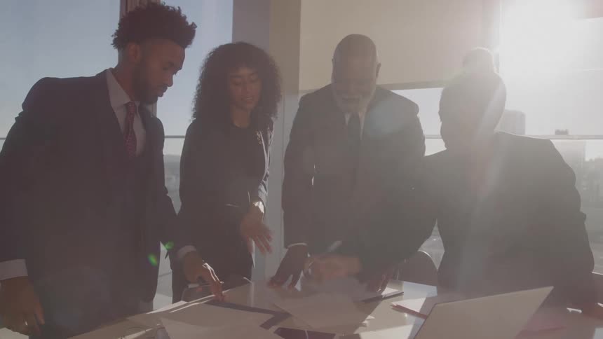In a sun drenched conference room a group of four diverse business professionals collaborates over documents spread across the table