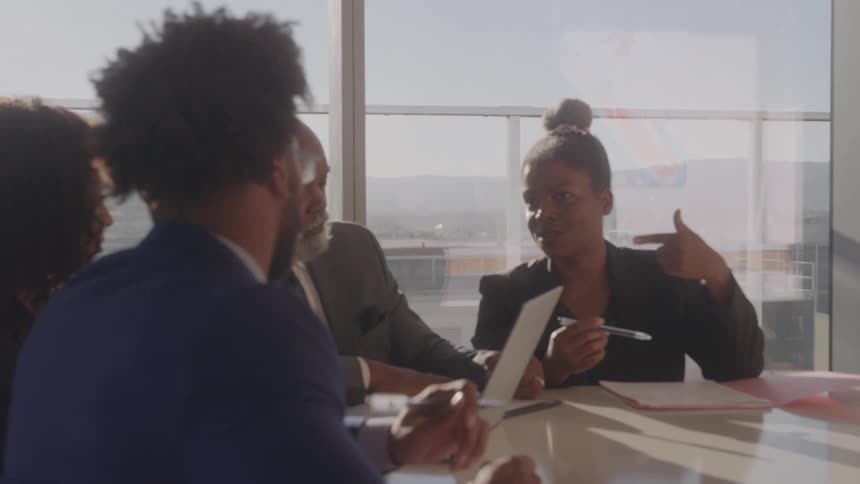 A woman sits at a table with a group of people and talks while gesturing with a pen
