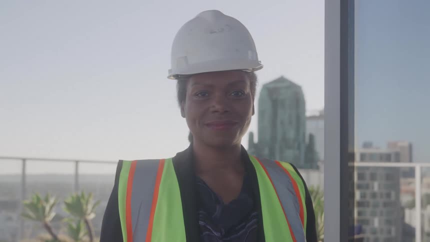 A woman wearing a hard hat and a reflective vest smiles at the camera with a city skyline in the background