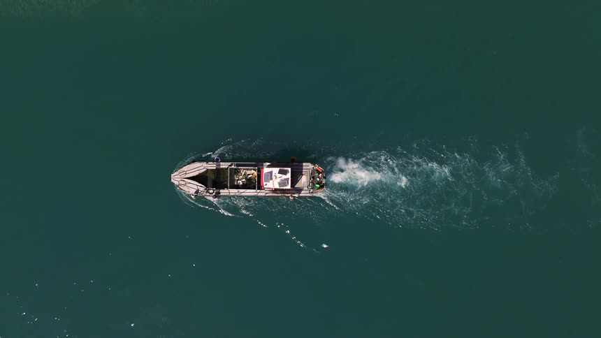 An aerial shot shows a small gray motorboat moving from left to right across a large body of deep green water the boat leaves a white bubbly wake behind it as it glides along