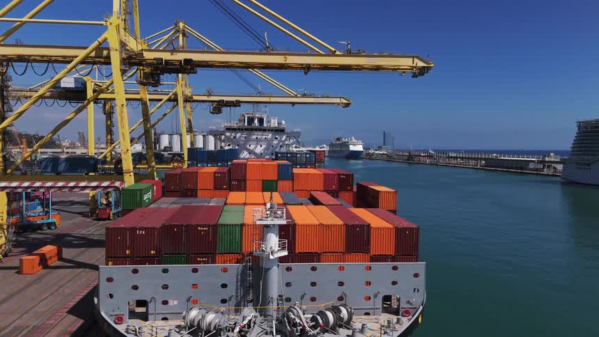 A cargo ship laden with a large stack of multicolored containers is docked at a busy port in the background large cranes loom over the water and other ships are visible in the distance