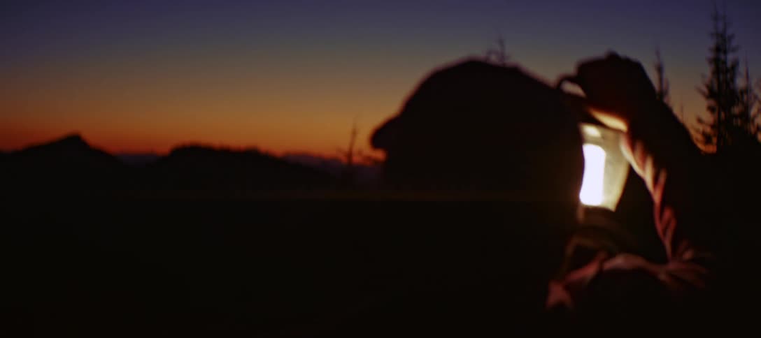 Man in a mountainous area at night looks around while holding a lantern before sitting down