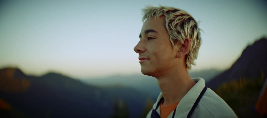 A medium closeup shot of a young man with blond hair looking out at the scenic view he has a warm smile on his face enjoying the beautiful landscape in the mountains