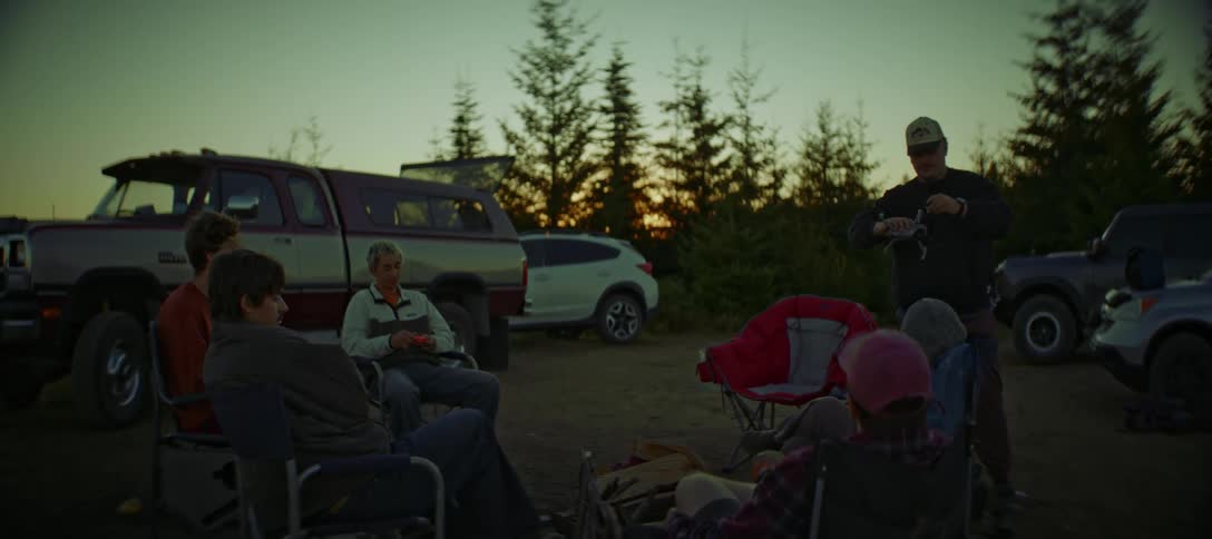 People sitting outdoors listen to a man holding a drone while red tints the horizon partially blocked by nearby conifers
