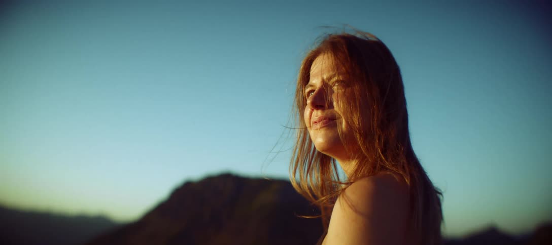 A side view of a woman with red hair looking out into the distance with a gentle breeze blowing through her hair the shot is set against a clear blue sky with the sun shining on her face