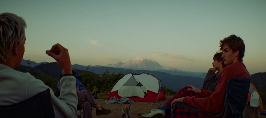 A group of friends sit around a fire pit at a campsite with a tent and a mountain range behind them