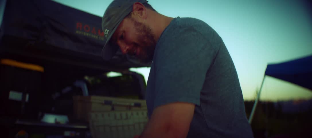 A man in a gray shirt looks down while camping outdoors at dusk