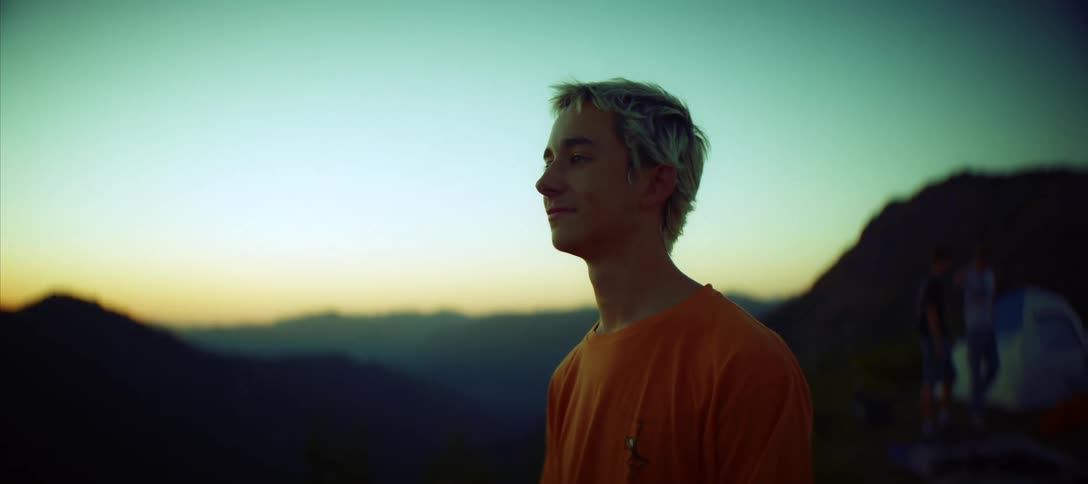 Blonde man stands looking at the scenery while he and his friends camp in mountainous terrain