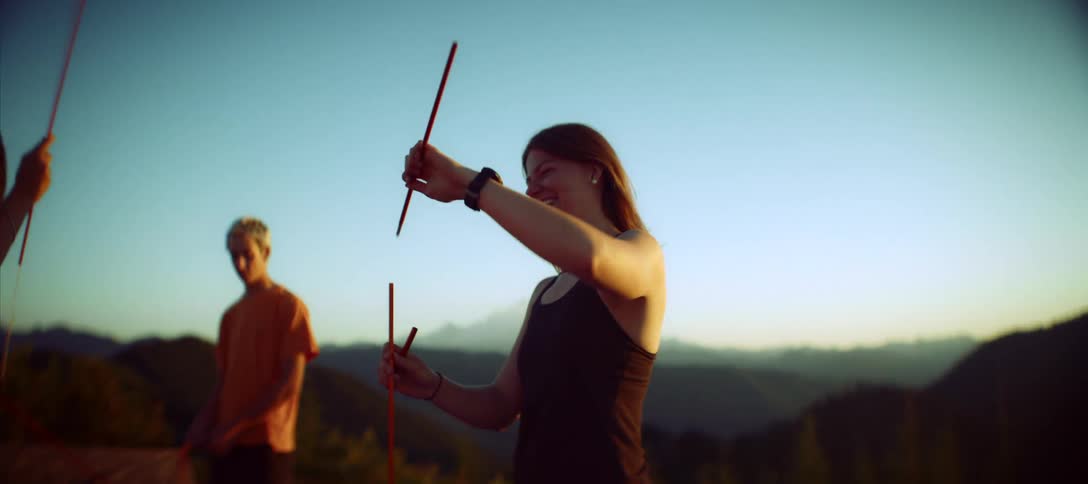 A smiling woman is shown in a closeup shot holding two poles for a tent in the background a man can be seen standing a few feet behind her they both appear to be setting up camp on a mountain top