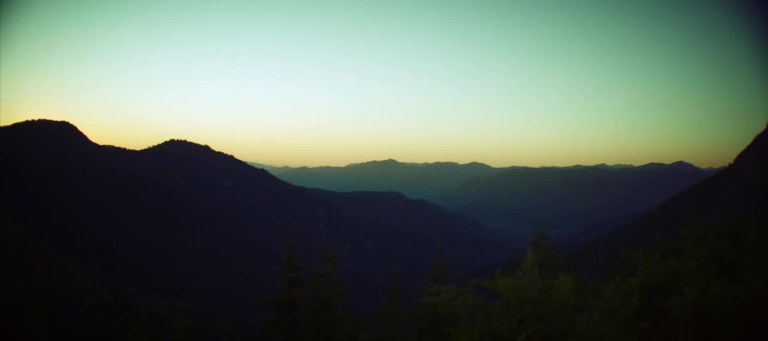 A view of a mountain range at sunset shows dark mountain silhouettes against a clear blue and gold sky