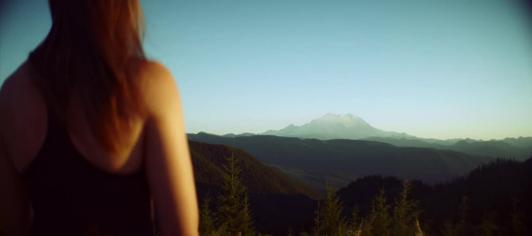 A view from behind of a woman with long red hair wearing a dark tank top looking out at a large foggy mountain range on a clear day with a forest in the foreground