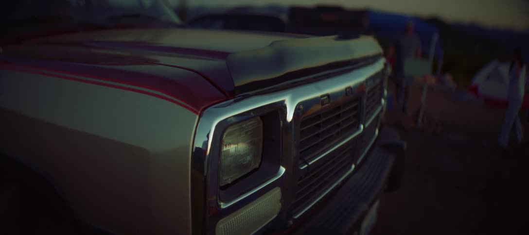 A shot of the front of a vehicle shows the hood grille and headlight of a pickup truck and people in a campsite on the background