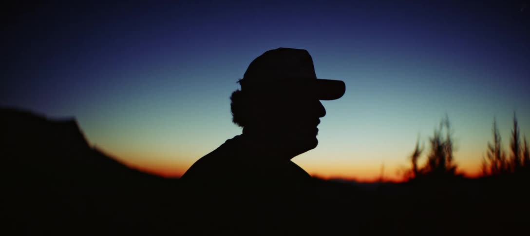A closeup backlit shot of a person in a cap and shirt with the light creating a silhouette of their head and shoulders the person appears to be watching a sunrise or sunset and the horizon behind them is cast in warm hues of orange and yellow