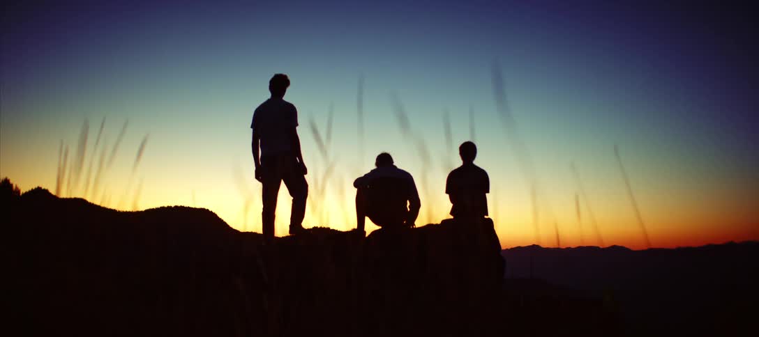 A low view of the silhouettes of three friends standing and sitting on a rocky hill as they watch the sun set over a distant mountain range
