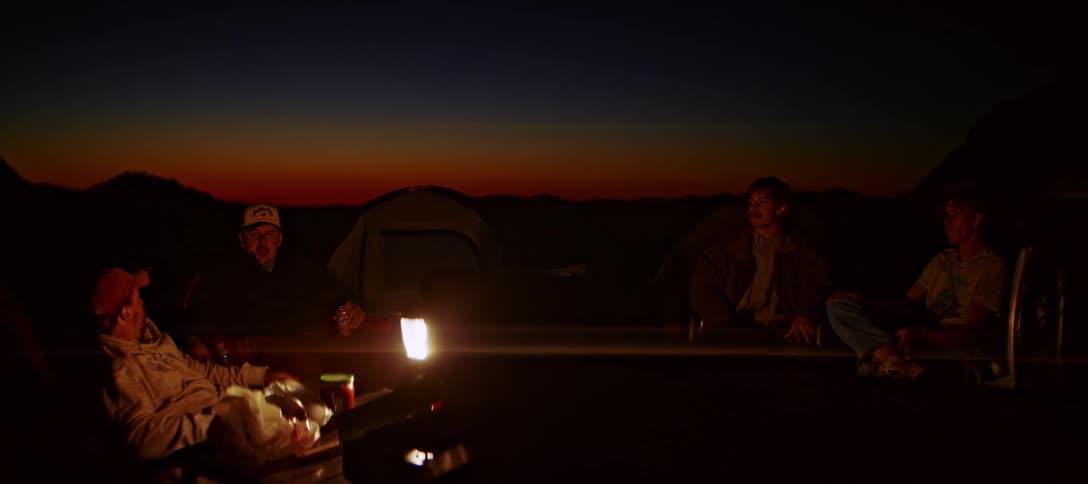 A group of friends sit around a lantern at their campsite at sunset