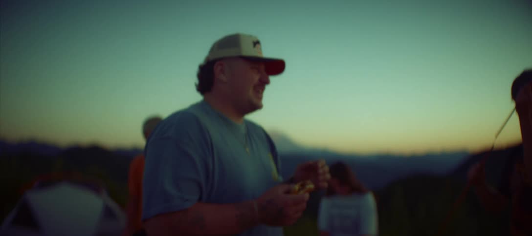 A group of people with a blurry figure in the foreground stand outside talking and laughing the shot is set against the backdrop of a mountain range at dusk and the sky is filled with tones of blue and orange
