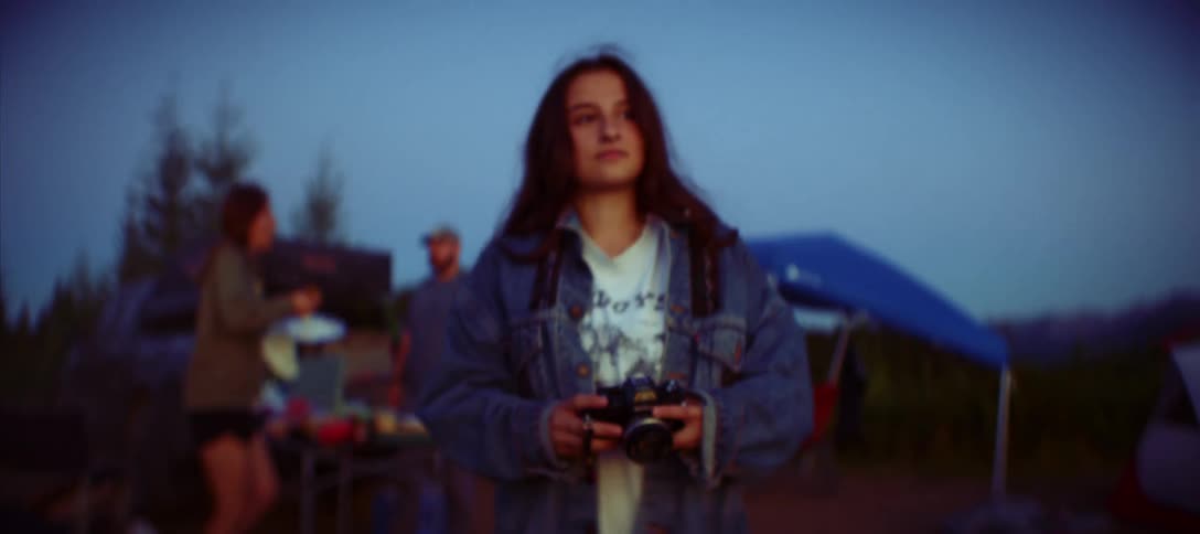 A woman is standing at a campsite holding a camera and bringing it up to her face to take a photograph while other people are in a campsite