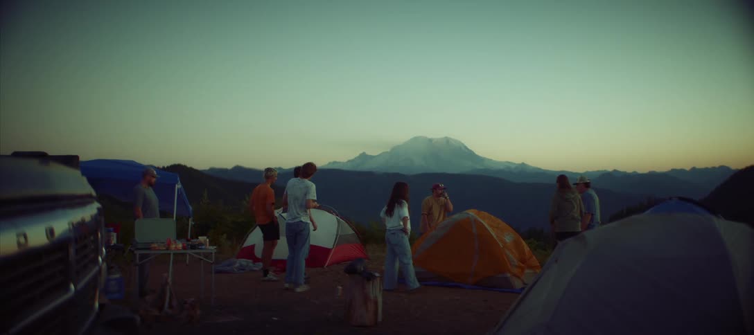A group of young adult friends socialize and set up their tents at a scenic mountain campsite as the sun rises or sets behind a large snowcapped peak