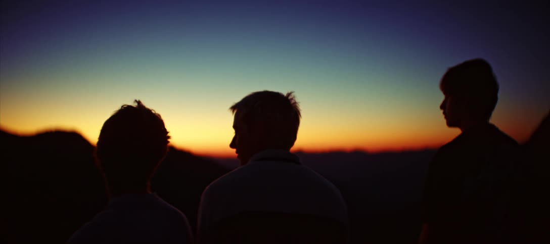 A long view of the silhouettes of three friends standing and watching the sun set over a distant mountain range with a beautiful colorful and peaceful sky