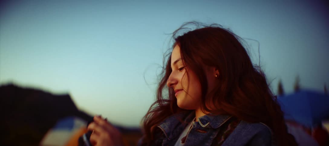 A close profile view shows a young woman with long brown hair wearing a jean jacket and taking a photo with a black camera at a campsite