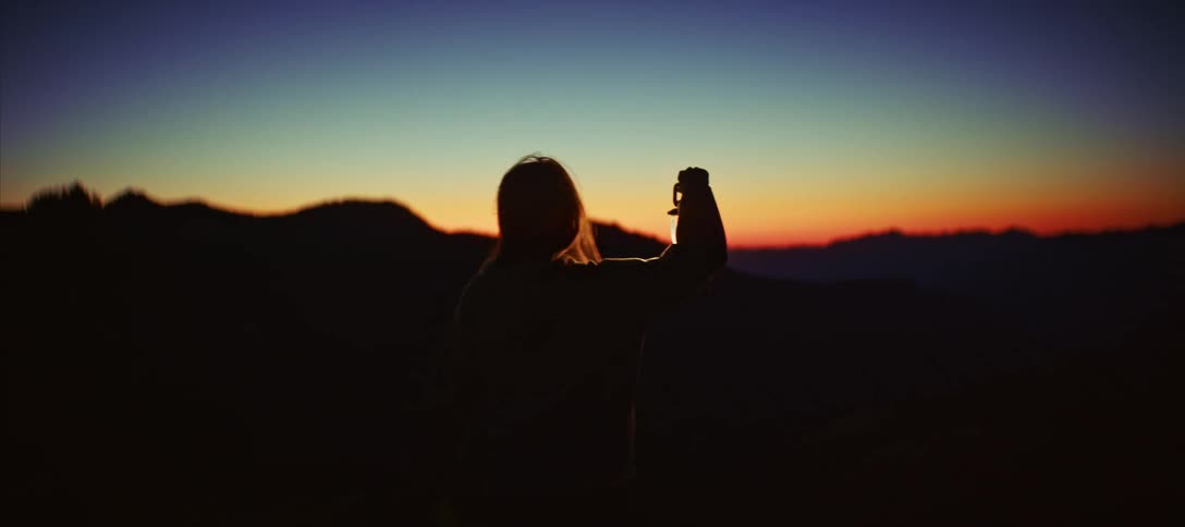 A silhouetted person with long hair holds a glowing lantern up in the air while standing on a mountain ridge and watching the sun set over a distant landscape