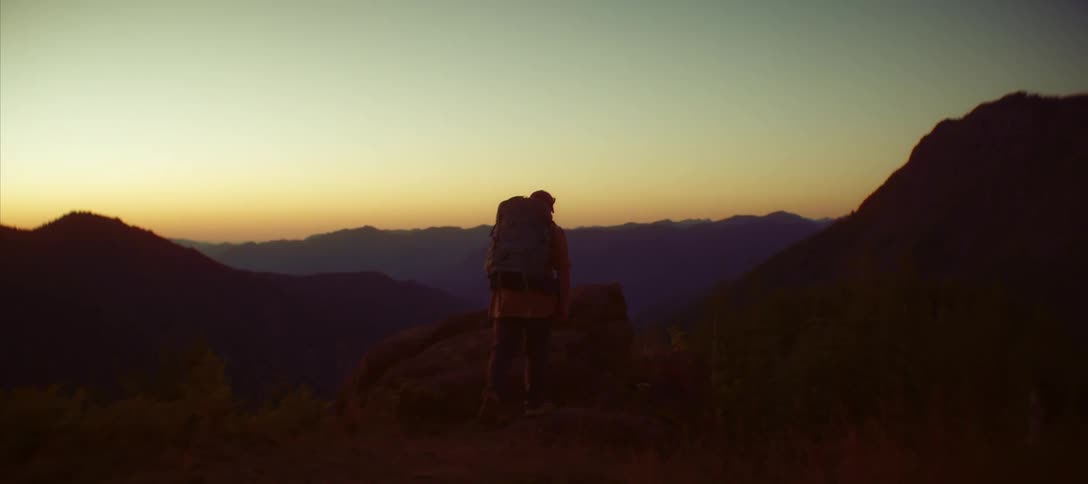 Male backpacker hiking through mountainous terrain climbs a boulder to view the scenery while red tints the horizon