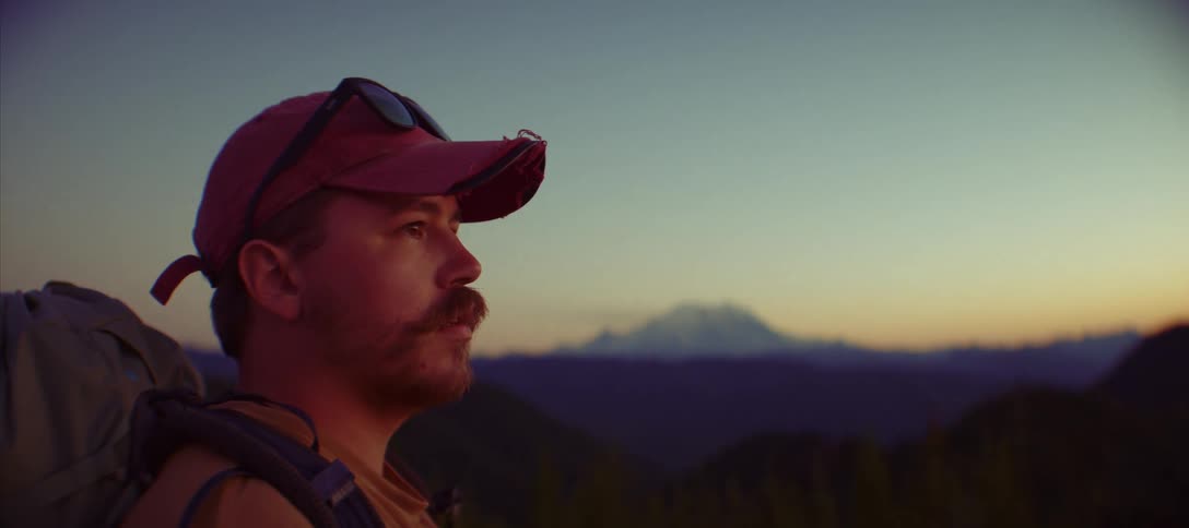 A man with a mustache and a hat looks out from a mountain ridge at a beautiful landscape with a large foggy mountain in the background at sunset