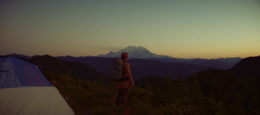 A man wearing a backpack stands on a mountain top watching the sunrise the camera focuses on the man in the foreground with a tent barely visible in the left corner while a snowcovered mountain is visible in the distance