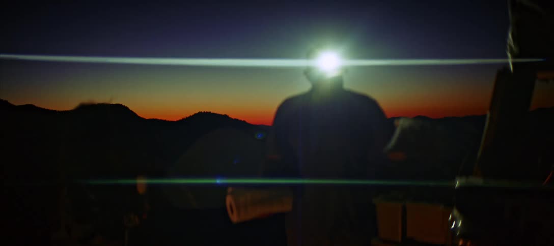 A silhouetted shot shows a man standing outdoors at night with the sky a beautiful gradient of orange yellow and blue at dusk he is wearing a headlamp which he turns on and off as he speaks to someone offscreen