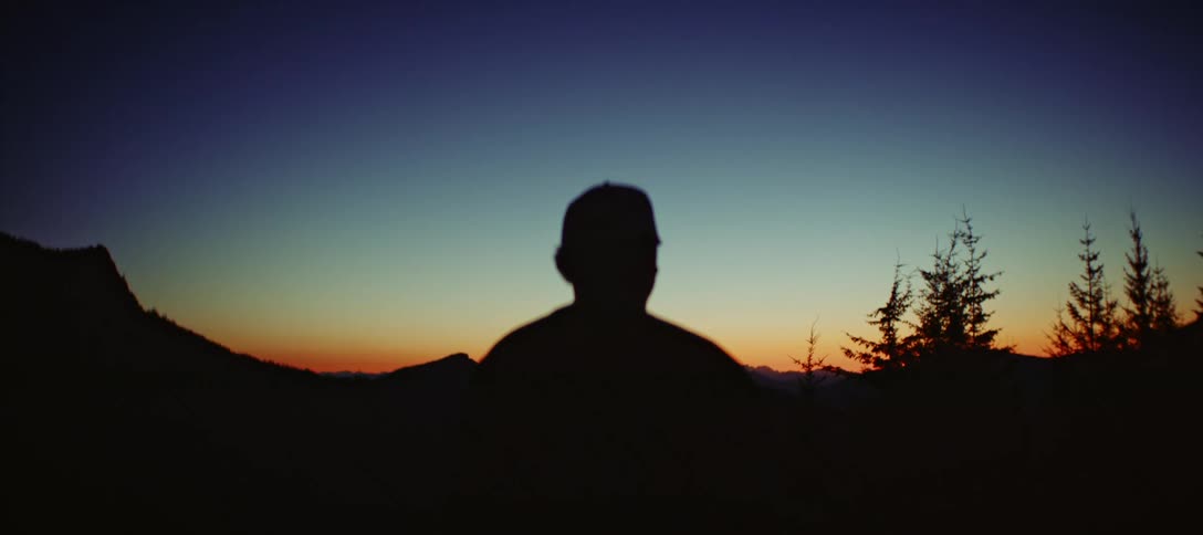 A closeup backlit shot of a person in a cap and shirt with the light creating a silhouette of their head and shoulders the person appears to be watching a sunset or sunrise and the horizon behind them is a vibrant blend of orange and yellow