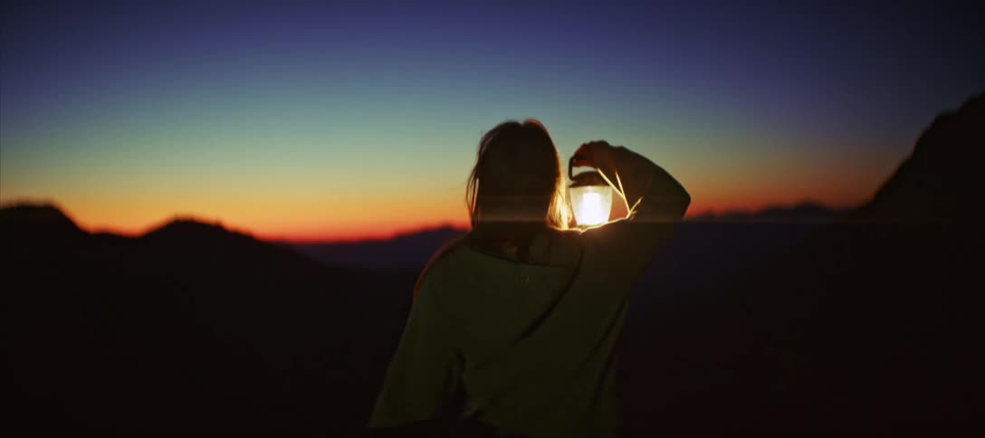 Woman holding a lantern and walking through a natural area with a landscape of mountains with a reddish sky on the horizon