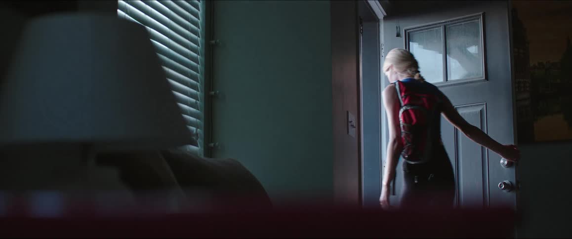 A side view of a young woman in a tank top and leggings is shown from a dark corner as she opens the door to a bedroom with light coming through the blinds