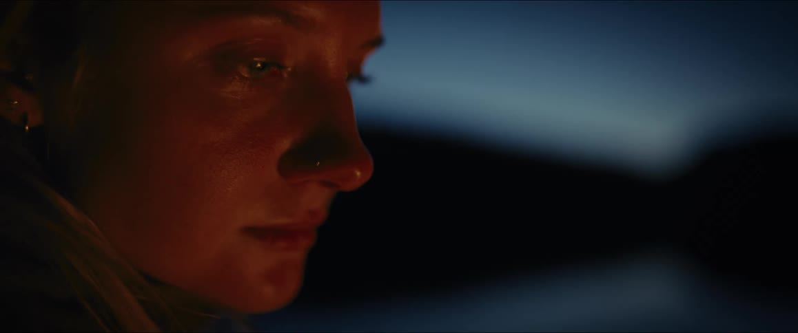 A close side view of a womans face shows her looking down with a serious expression with a faint warm glow on her skin at night